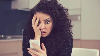Mladá žena se zoufalým a zděšeným výrazem kouká na mobilní telefon, který má v ruce. Druhou rukou si zakrývá obličej