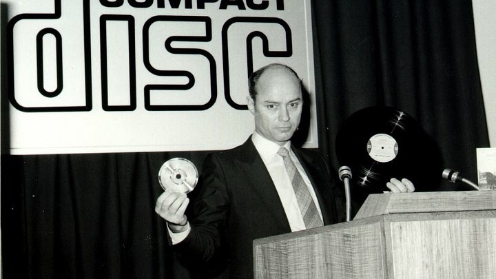 Výzkumem a vývojem nových audio disků, využívajících laserovou technologii, které by konkurovaly gramofonovým deskám, se ve firmě Philips začali zabývat v 70. letech. Tehdejší technický ředitel firmy Lou Ottens navrhoval, aby nový audio disk byl menší než vinylová deska a aby pojmul hodinový záznam. V roce 1977 se práce na vývoji zintenzivnily a začal se hledat pro disk vhodný název. Zvažovalo se například označení MiniRack, MiniDisc či CompactRack. Nakonec zvítězil název CompactDisc. Představitelé firmy se totiž domnívali, že by tento název mohl lidem evokovat úspěch audiokazet (Compact Cassette).
Na snímku šéf laboratoří společnosti Philips Joop Sinjou představuje v roce 1979 veřejnosti první kompaktní disk. Tehdy měl ještě průměr 11,5 cm a vešla se na něj hodina záznamu.